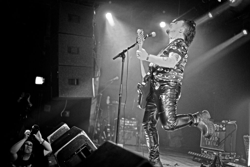 A photo of Jack Armando of Panic Priest. The photo is edited into black and white, and is taken from the right side of the stage. Mr. Armando is standing on stage, facing the crowd, playing his guitar and singing into the mic. He has his head thrown back and his left leg kicked up and back behind him, giving him a dramatic look.