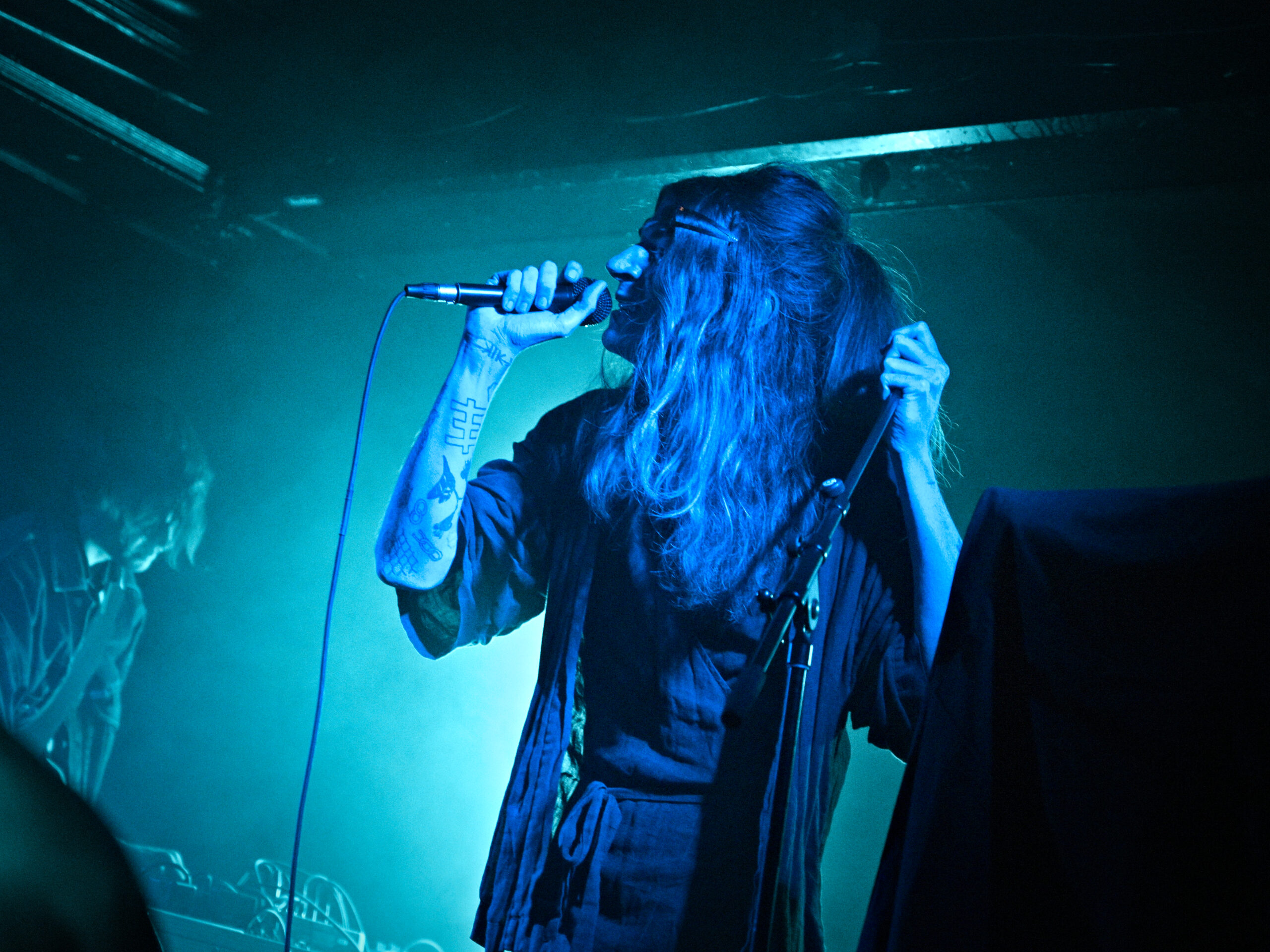 A photograph by Dancing Stag Photography. Both members of Korine are shown, one in the foreground, just off-center to the right, lit in dark blue, the other softly in the background to the left.